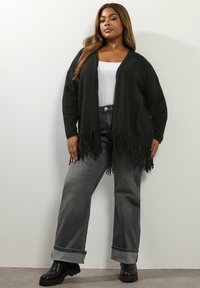 Kvinna i svart fransad cardigan, vit topp, grå uppvikta jeans och svarta stövlar, står mot en vit vägg med händerna vid sidorna.