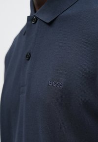 Tengerészkék galléros póló két fekete gombbal és a mellkason hímzett "BOSS" logóval, egy személy által viselve.