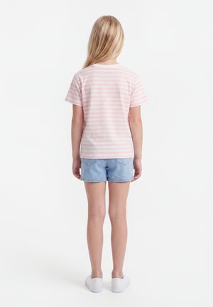 Enfant aux longs cheveux blonds portant un t-shirt rayé rose et blanc, un short en jean bleu clair et des chaussures blanches, debout dos à la caméra.