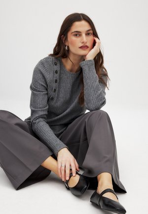 Pelēks ribota džemperis ar pogu detaļām uz pleca, apvienots ar brīviem, tumši pelēkiem platām biksēm un melniem dzīvokļiem, sēžot uz gaiša fona.