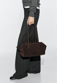 Person trägt grauen Pullover und schwarze weite Hosen und hält eine dunkelbraune Wildlederhandtasche mit langen Trägern.