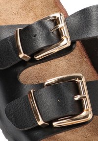 Las sandalias de cuero negro presentan dos correas ajustables con hebillas doradas, reposando sobre una plantilla texturizada de color marrón.