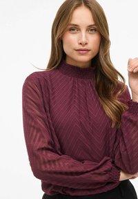 Blusa burdeos con un patrón de chevron texturado, cuello smock alto y mangas largas transparentes recogidas en los puños.