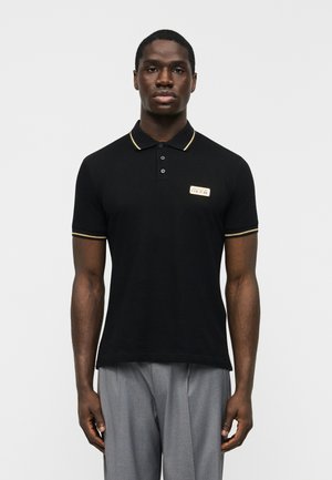 Mees, kes kannab musta polo särki kuldsete ääriste ja Versace Jeans Couture logoga, hallide pükstega, seistes valge tausta ees.