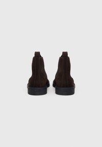 Botas de tobillo de ante marrón con paneles laterales elásticos, suelas de goma y costuras en contraste. Tacón bajo y marca visible en la parte trasera.