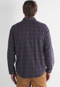 Homme portant une chemise à carreaux violette sombre à manches longues et un pantalon marron, se tenant de dos à la caméra devant un fond uni.