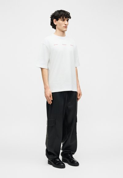 Άνδρας στέκεται φορώντας λευκό υπερμέγεθες T-shirt με μικρό κόκκινο κείμενο και φαρδιά μαύρη παντελόνια cargo με μαύρα παπούτσια.