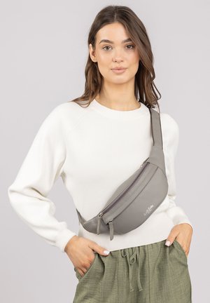 Graue Kunstleder-Schultertasche mit einem geschwungenen Design, Reißverschluss und verstellbarem Riemen, getragen über einem weißen Pullover und grünen Hosen.