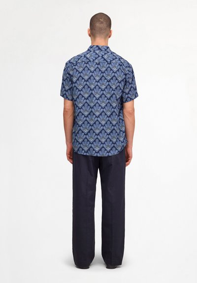 Homme de dos portant une chemise bleue à manches courtes avec un motif géométrique et un pantalon large bleu marine foncé sur fond uni.