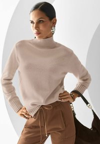 Frau in beigem Rollkragenpullover und braunen Jogginghosen posiert mit einer Hand auf der Hüfte und einer schwarzen Handtasche am Handgelenk, vor weißem Hintergrund.