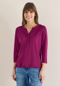 Femme souriante portant une chemise violette à manches longues avec des boutons et un jean bleu, debout devant un fond uni beige clair.
