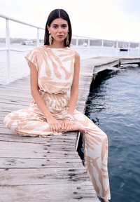 Combinaison beige clair avec des rayures crémeuses abstraites, fabriquée dans un tissu texturé. Dotée d'un décolleté large et d'une taille élastique.