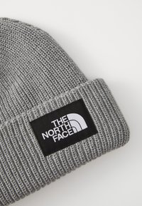 Grå strikket hue med ribbet tekstur, der har en sort lap med et hvidt logo. Stoffet ser blødt og strækbart ud.