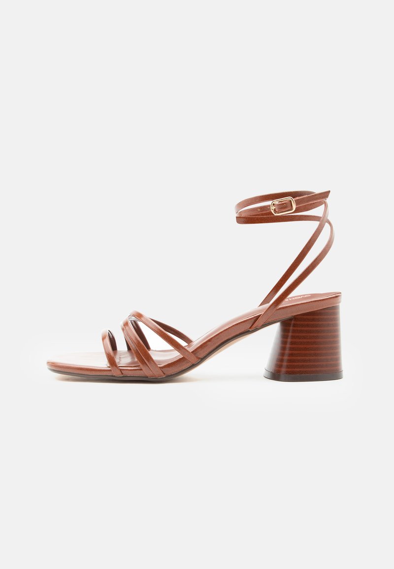 Sandale marron à brides avec talon bloc et boucle à la cheville, design minimaliste, texture en cuir lisse, vue de côté sur fond uni.