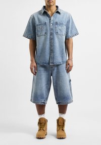 Jasnoniebieska, krótkim rękawem koszula dżinsowa oraz szorty do kolan z dwiema kieszeniami na piersi, ozdobione subtelnymi haftami. Noszone z brązowymi butami roboczymi.