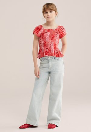 Fiatal lány piros mintás rövid ujjú blúzban, világoskék bő szárú farmerben és piros szövött bebújós cipőben, kezét zsebében tartva áll.