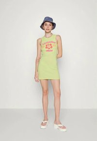 Vestido verde sem mangas com um design justo, apresentando um padrão floral em rosa e amarelo. Combinado com um chapéu de balde azul e sandálias brancas.