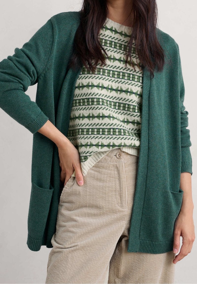 Femme portant un cardigan vert ouvert à l’avant, un pull vert et blanc à motifs et un pantalon en velours côtelé beige, main reposant dans la poche.