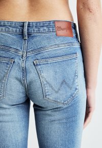 Jeans de denim azul claro con una etiqueta de cuero con la marca "Wrangler", dos bolsillos traseros con un patrón de costura en zigzag.