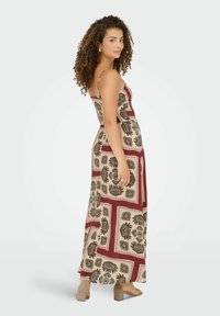 Femme aux cheveux bouclés portant une robe maxi sans manches à motifs beige et bordeaux et des sandals à talons carrés beiges, se tenant de profil.