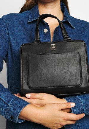 Mujer con camisa de mezclilla azul que sostiene un bolso de cuero negro con doble asa y bolsillo delantero, mostrando el logotipo de la marca.