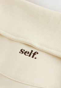 Tessuto color crema con colletto a coste. La parola "self." è ricamata in marrone vicino alla cucitura del colletto. Texture morbida, design minimale.