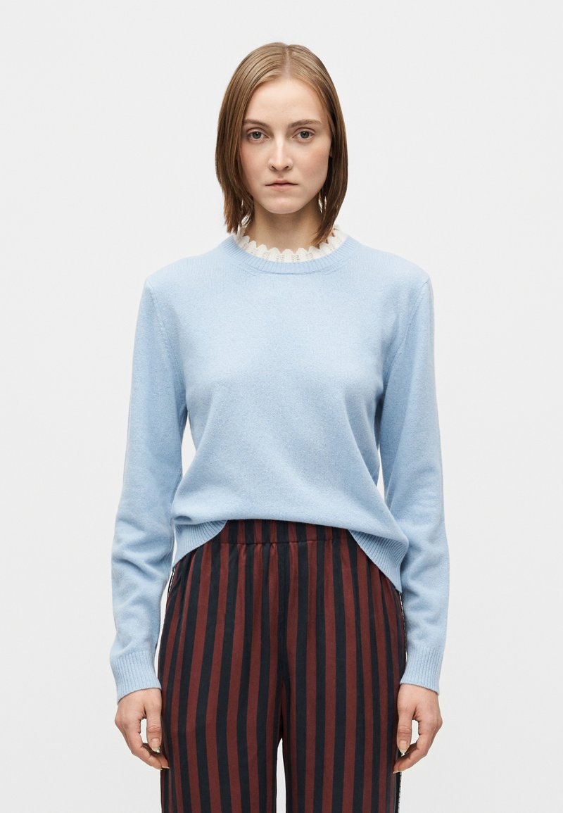 Hellblauer Pullover mit geripptem Ausschnitt und gewelltem Detail, kombiniert mit rot-schwarz gestreifter, vertikal gestreifter Hose. Glatte Textur, figurbetontes Design.