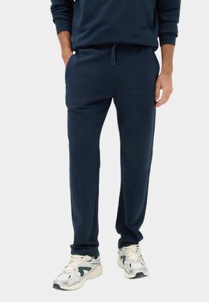 Uomo con pantaloni della tuta blu navy e sneaker bianche in piedi su sfondo bianco con le mani parzialmente nelle tasche.