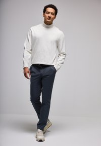 Suéter de cuello alto blanco y texturizado, combinado con unos pantalones oscuros de cintura elástica. Zapatillas deportivas en tonos neutros completan el atuendo.