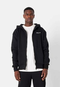 Sudadera negra con cremallera hecha de tela suave, cuenta con capucha ajustable, bolsillos frontales y un pequeño logo en el pecho. Usada sobre una camisa blanca.