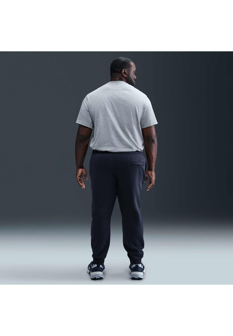 Uomo in piedi di spalle alla fotocamera, indossa una maglietta grigio chiaro, pantaloni scuri e scarpe da ginnastica, su uno sfondo scuro e uniforme.