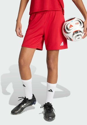 Person trägt rote Sportshorts, weiße Adidas-Socken, schwarze Fußballschuhe und hält einen weißen und roten Adidas-Fußball neben sich.