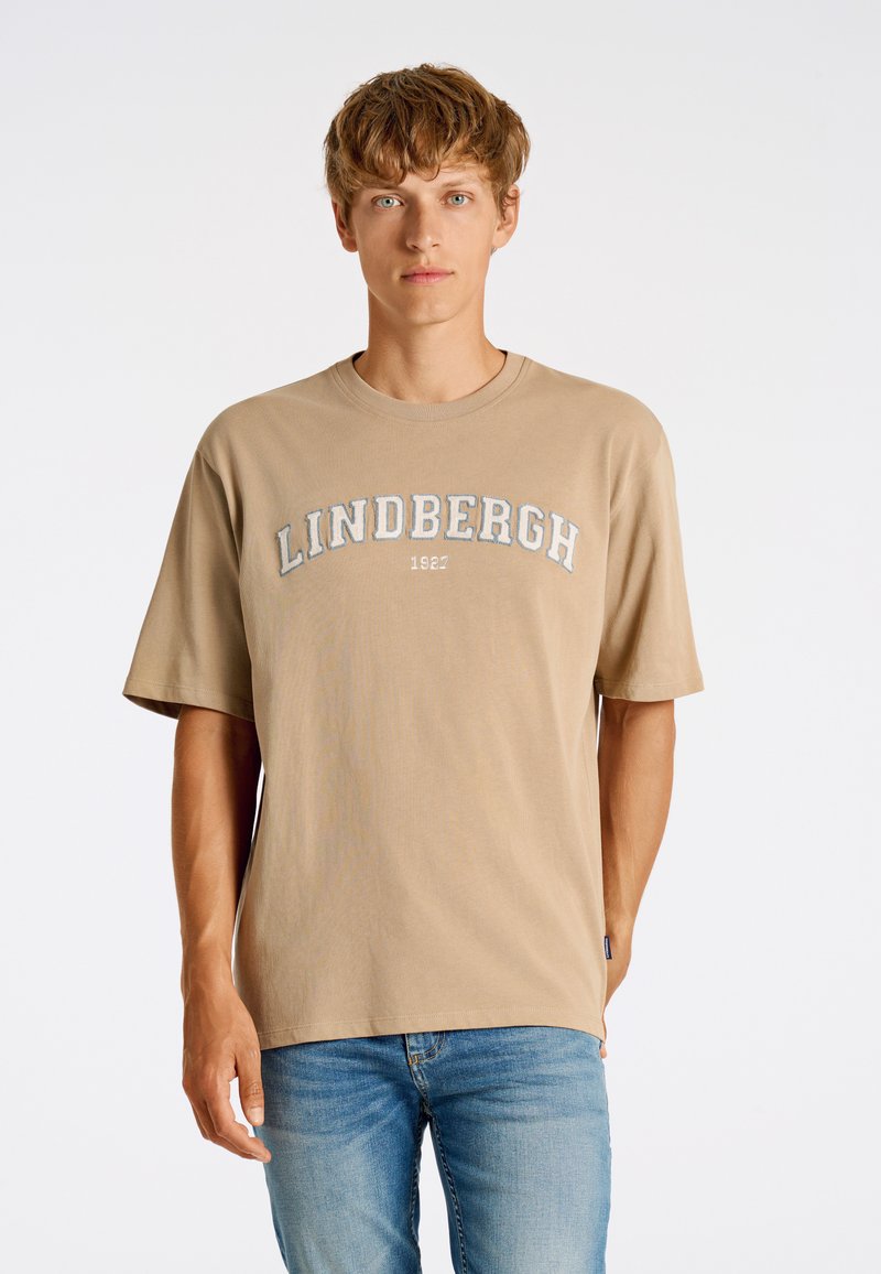 Beige puuvillapaita, jossa on lyhyet hihat, pyöreä pääntie ja kontrastivärinen kirjailtu teksti "LINDBERGH 1927". Yhdistetty vaaleansinisiin farkkuihin.