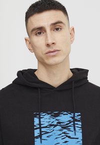 Felpa nera con un graffito blu di onde d'acqua. Realizzata in un materiale morbido, presenta un cappuccio con cordino e una vestibilità comoda.