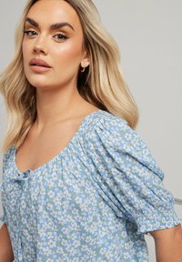 Blusa floreale azzurro chiaro con maniche a sbuffo e scollatura quadrata, caratterizzata da un dettaglio annodato nella parte anteriore e piccoli motivi floreali bianchi e verdi.