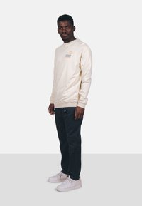 Camisa de manga larga crema con un pequeño gráfico naranja y líneas azules onduladas en el pecho, combinada con pantalones oscuros y zapatillas blancas.
