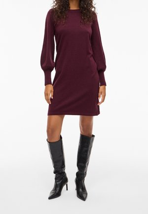 Femme portant une robe en maille bordeaux à manches longues bouffantes, chaussant des bottes noires à talons, pointues et montantes jusqu'au genou.
