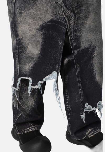 Dunkelblaue Jeans mit zerstörten Stellen, ausgefransten Säumen und verblassten Patches. Sie haben einen lockeren Schnitt und einzigartige Verblassungsmuster. Schwarzes Schuhwerk ist sichtbar.