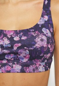 Bralette com estampado floral e decote arredondado, apresentando tons de roxo e rosa sobre um fundo escuro, feito de um tecido macio e elástico.