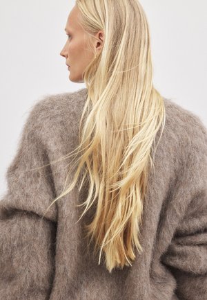 Mäkký hnedý mohair sveter s nadrozmerným siluetou; má chlpatú textúru a spadnuté ramená, nosený s dlhými, rovnými blond vlasmi.