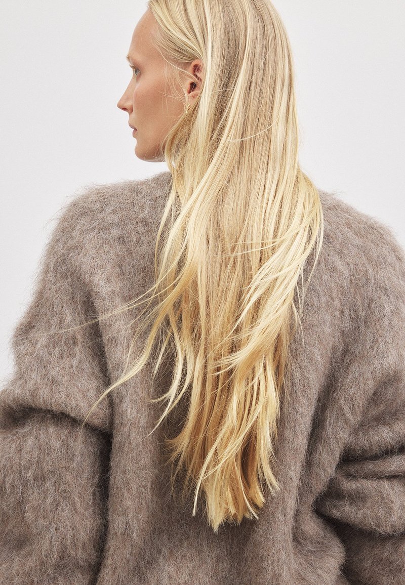 Miękki brązowy sweter z moheru o oversize'owej sylwetce; charakteryzuje się puszystą fakturą i opadającymi ramionami, noszony z długimi, prostymi blond włosami.