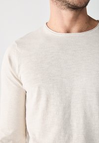 T-shirt à manches longues de couleur beige clair encolure crew, fabriqué dans un tissu doux et texturé, présentant une finition lisse sans motifs ni coutures visibles.
