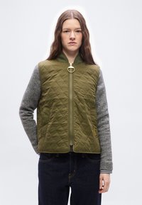 Νεαρή γυναίκα που φοράει γιλέκο χακί με ραφές και χρυσλό φερμουάρ πάνω από γκρι πλεκτά μανίκια και σκούρα τζιν, κοιτάζοντας εμπρός με ουδέτερη έκφραση.