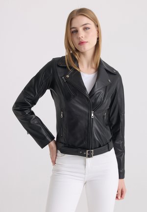 Donna con capelli biondi lisci che indossa una giacca di pelle nera sopra una camicia bianca e pantaloni bianchi, in piedi contro uno sfondo chiaro.