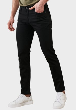 Mężczyzna w czarnych jeansach o wąskim kroju i białych trampkach, z jedną ręką wsuniętą do przedniej kieszeni, stojący na tle jednolitego tła.