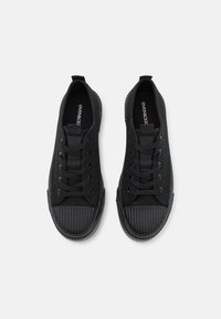 Czarne płócienne sneakersy z chropowatą gumową osłoną na palce i grubymi sznurowadłami. Posiadają płaską podeszwę i pętelkę z tyłu ułatwiającą zakładanie.