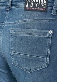 Jean en denim bleu avec une poche arrière dotée d'un bouton argenté, de détails cousus et d'une étiquette en tissu avec un texte coloré.