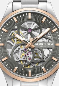 Automatyczny zegarek z przezroczystą szaro-skalową tarczą, różowozłotymi akcentami, srebrnymi elementami i skomplikowanymi szczegółami mechanicznymi.