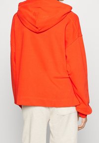 Felpa con cappuccio arancione, con spalle scese, vestibilità comoda e una texture del tessuto morbida. Presenta una tasca a marsupio e polsini a costine.