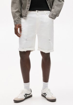 White denim shorts with distressed details and rolled hems. Worn with textured gray jacket, white socks, and white sneakers with black accents.